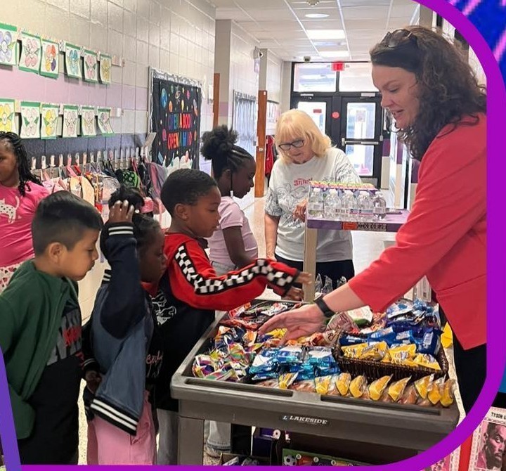 Mrs. Thompson is standing by a cart filled with treats like cookies and chips.  A group of students are standing at the cart looking at the treats.