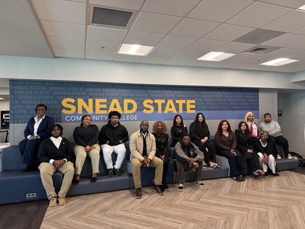 Pictured and seated from left to right is Ms. Holman in dark blue suit with white shirt three students in black tops, beige and white pants, Mr,. Steger with tan jacket, dark shirt and khaki pants, student with black jacket with buttons, Mr. King with dark jacket and dark green pants, 6 girl students next to him with dark clothin g and boy student on the end in a grey jacket and dark pants