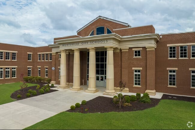 Front entrance of Lee High School in Huntsville, Alabama, showing the brick building with tall cream-colored columns, large front windows, landscaped shrubs, and a sidewalk leading to the main doors under the “Huntsville City Schools” sign.