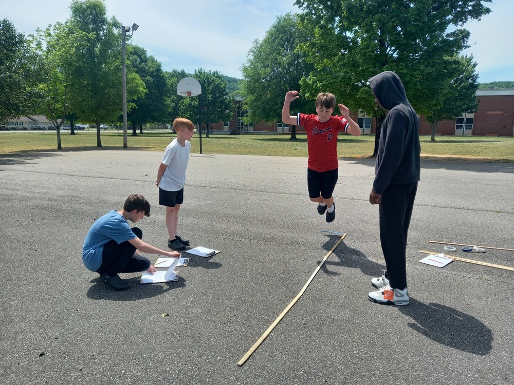 one student in a red shirt is jumping while three students stand around him to measure how far he jumps