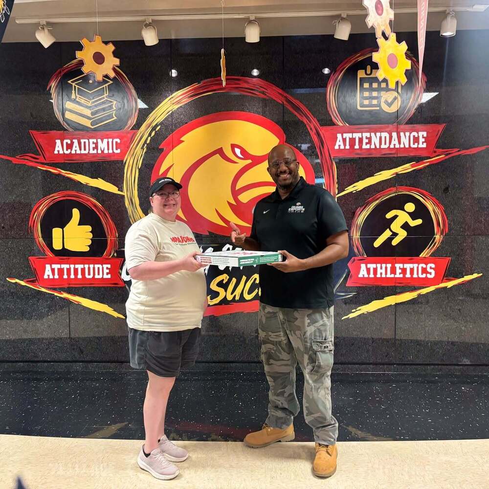 On the left is Papa John's Pizza employee in yellow shirt and red writing with dark shorts handing Mr. Merriweather a pizza box who is standing on the right with a black shirt and army pants