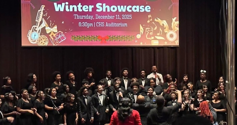 Pic of the Winter Showcase Sign above the stage with the choir dressed in black and audience in the seats