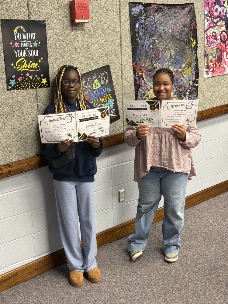 Two students proudly display spelling bee certificate in front of a wall, showcasing their achievements