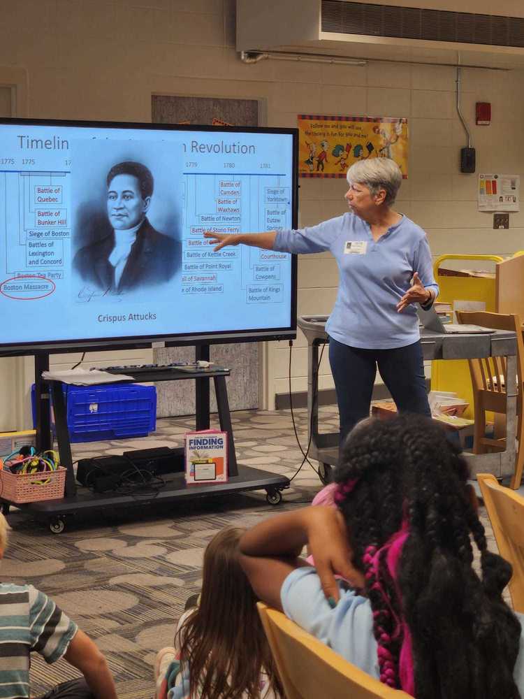 A member of the Daughters of the American Revolution points to a slide about Crispus Attucks and the American Revolution while speaking to a group of attentive students. The students sit on the floor in a classroom filled with books and learning materials.