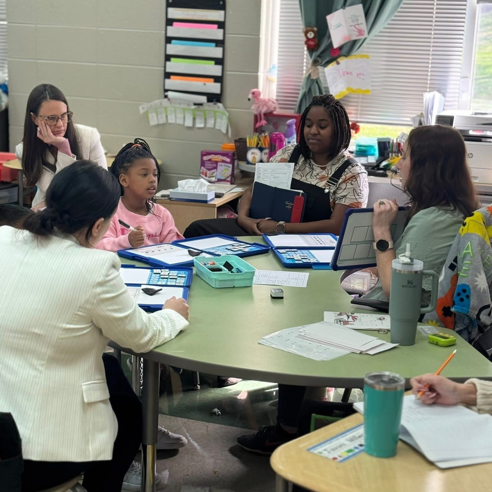 4 adults pictured sitting around the green table with a student in a pink shirt