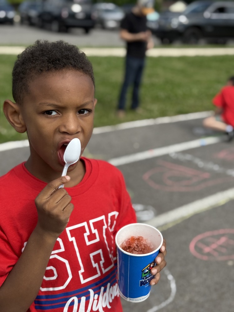 A young student wearing a red Sonnie Hereford Wildcats shirt enjoys a cup of Kona Ice while standing on the playground during an outdoor school celebration. Chalk drawings cover the pavement in the background as other students and staff participate in activities on a sunny day.