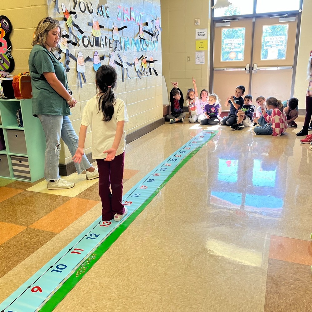 A first grade student walks beside her teacher on a giant number line in the hallway in front of her classmates.