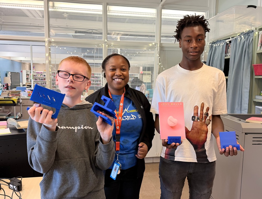 Two PLTW students and Ms. Moncur are in the library office showing off their newly created 3-D printer projects for the library.