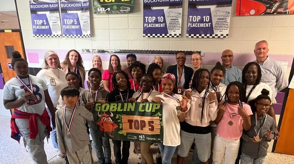 A group of students are standing in the hallway, along with administration from Dawson and the Central Office.  They are holding a banner that says "Goblin Cup Top 5".  On  the wall behind them are various banners hanging from previous Greenpower races.