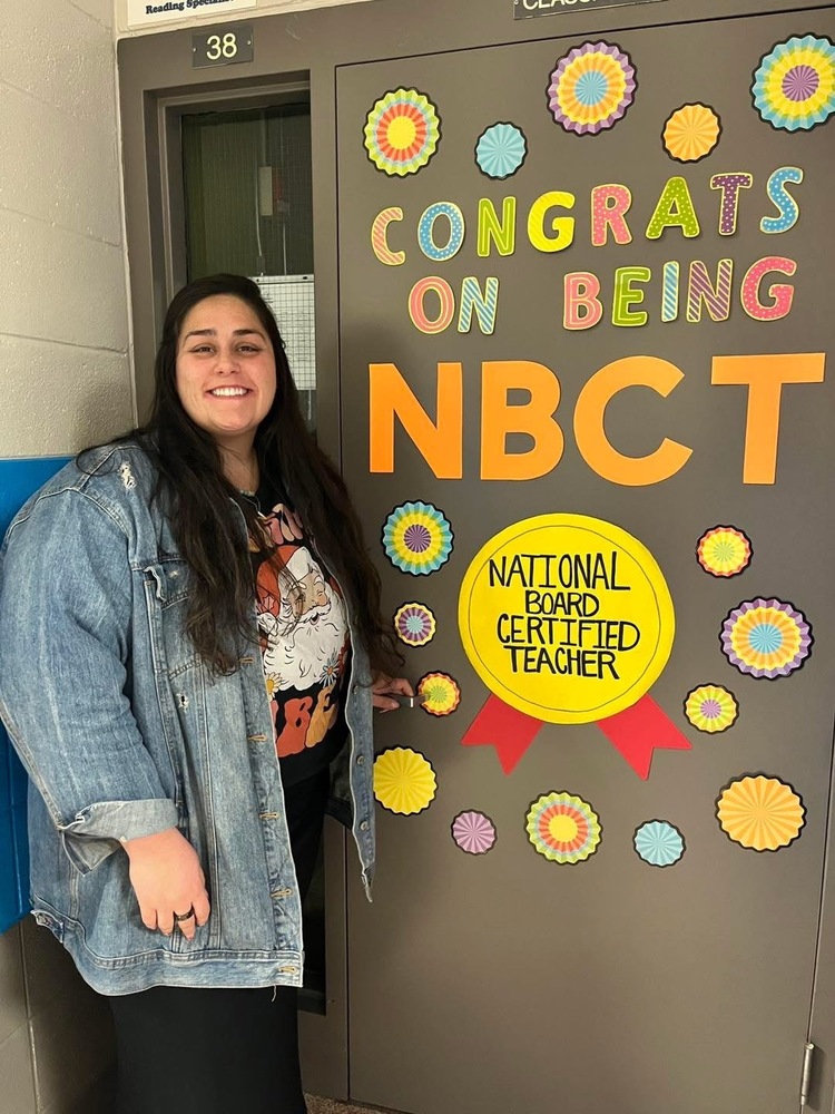 Ms. Hackett stands beside a decorated door celebrating National Board Certified Teacher achievement, with colorful paper flowers and congratulatory text.