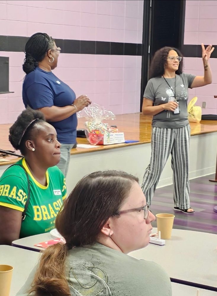 Four mothers are gathered in the cafeteria. Two are standing facing other parents. The rest are seated at a table.