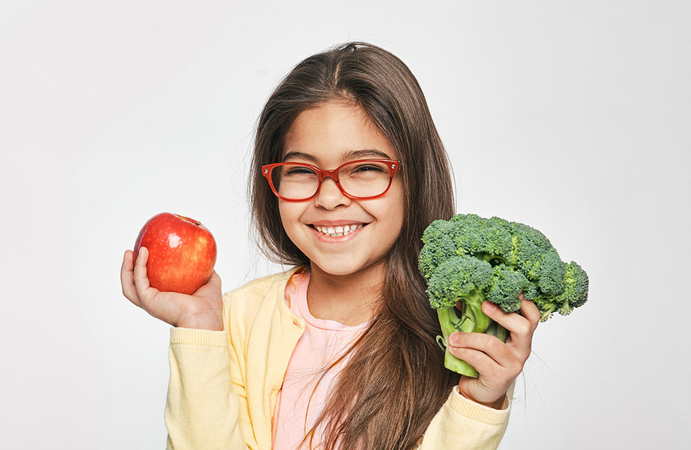 Girl with red glasses smiling and holding an apple and a head of broccoli