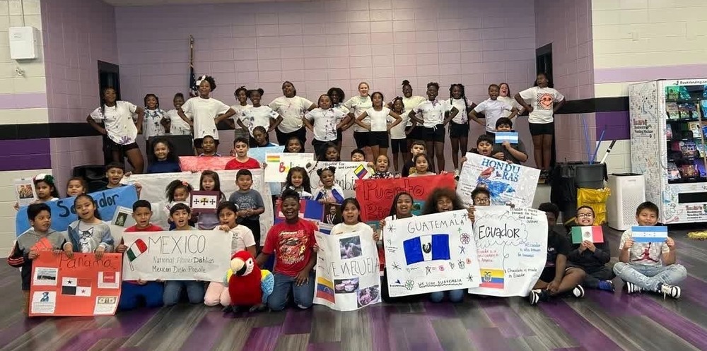 A large group of students are posed in the cafeteria . Students are holding posters from different countries like Mexico, Ecuador, and Guatemala.