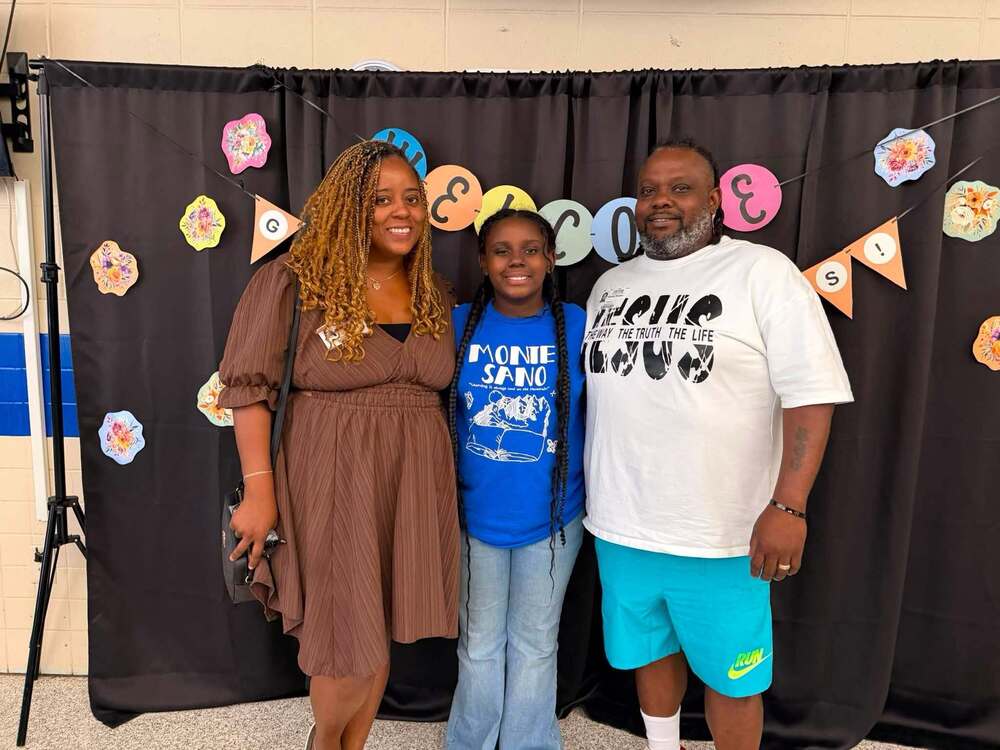 Student standing with two adults in front of photo backdrop that says welcome