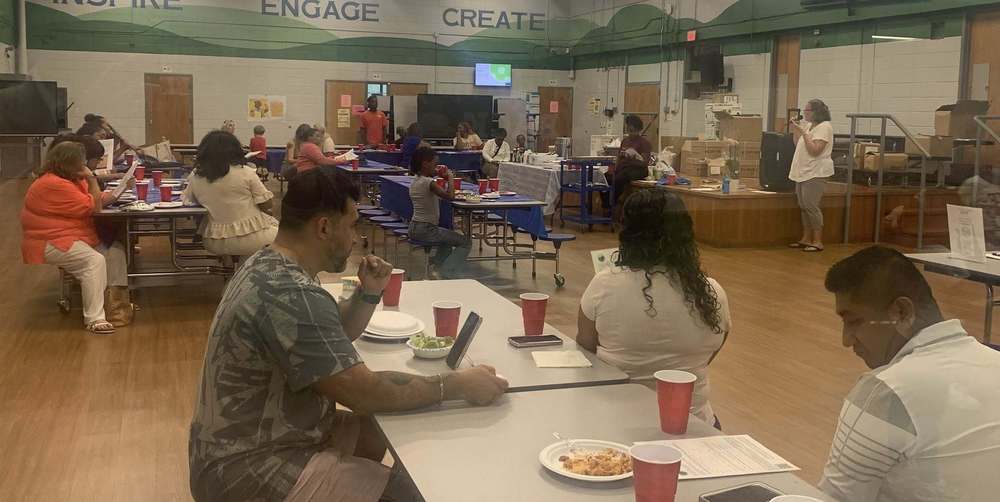 Families and staff sit at tables eating while PTA President Holly Jones speaks into a microphone at the front of the room.