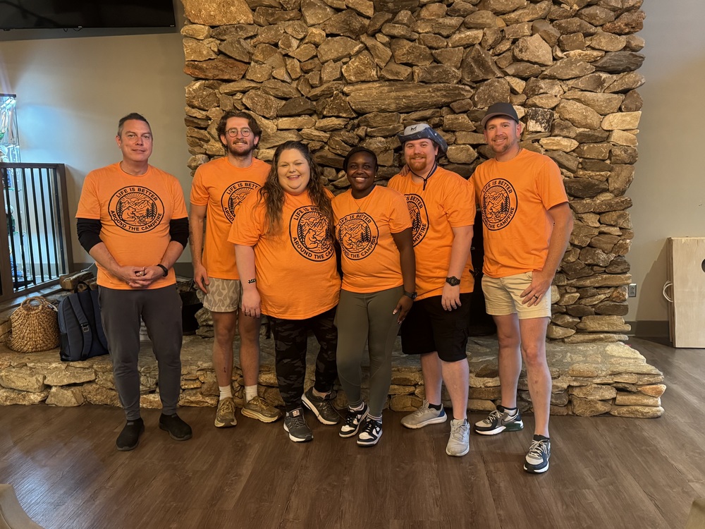 A group of teachers in orange shirts stands in front of a textured stone wall, smiling and posing for the photo.