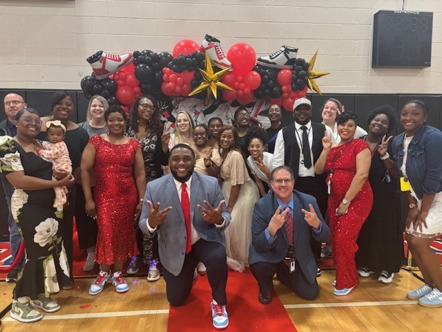 A large group of adults and children pose together in a school gym decorated with a red, black, and gold balloon arch featuring sneaker-themed accents. Two men kneel in front on a red carpet while others stand behind them smiling and making peace signs. Several people are dressed in red outfits and sneakers, and one person holds a small child, capturing a festive, celebratory atmosphere.