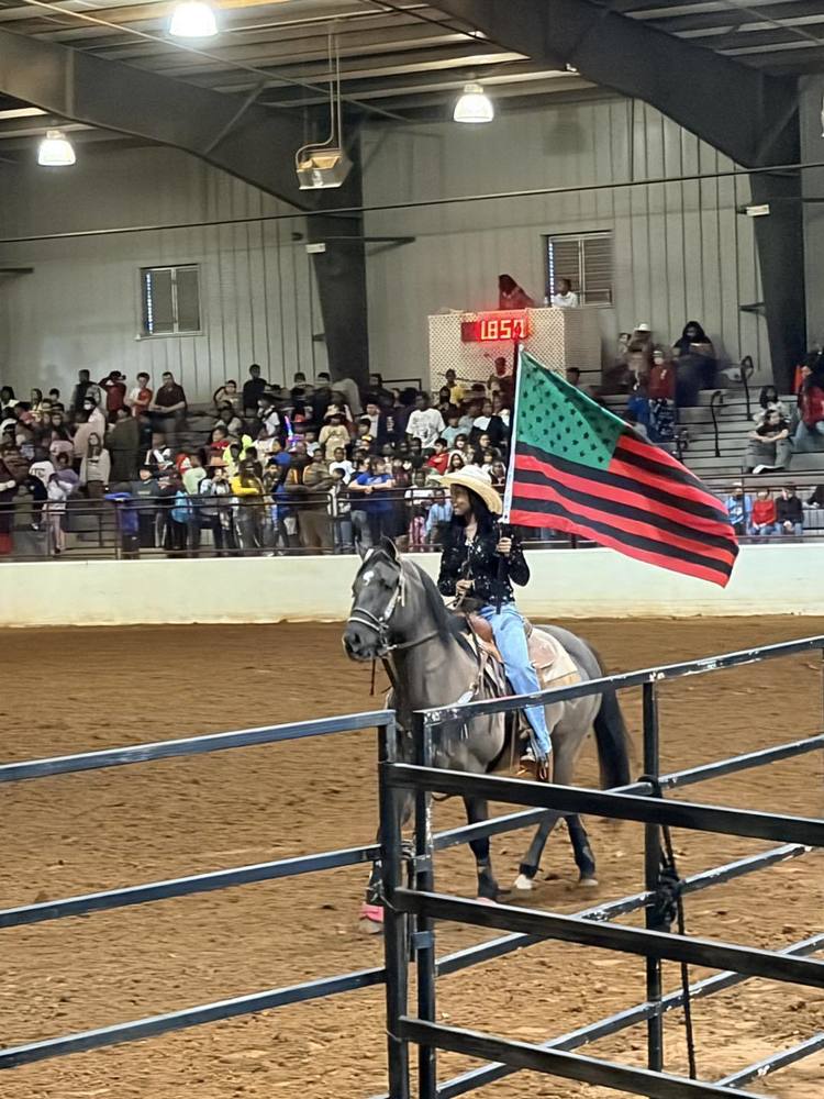 A cowgirl is riding a grey horse inside an arena full of students.  She is carrying the red, green, and black flag for Black History.