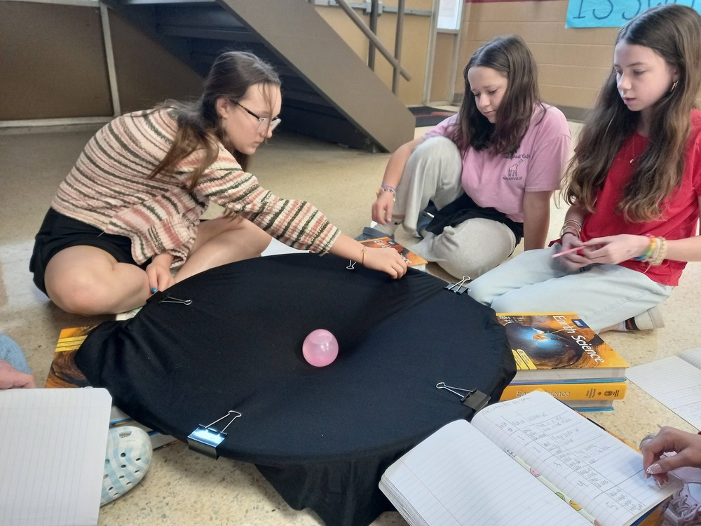 three 6th grade students are sitting around  a black circle and one student is  about to roll a marble on the edge to demonstrate the effects of gravity between two objects of mass