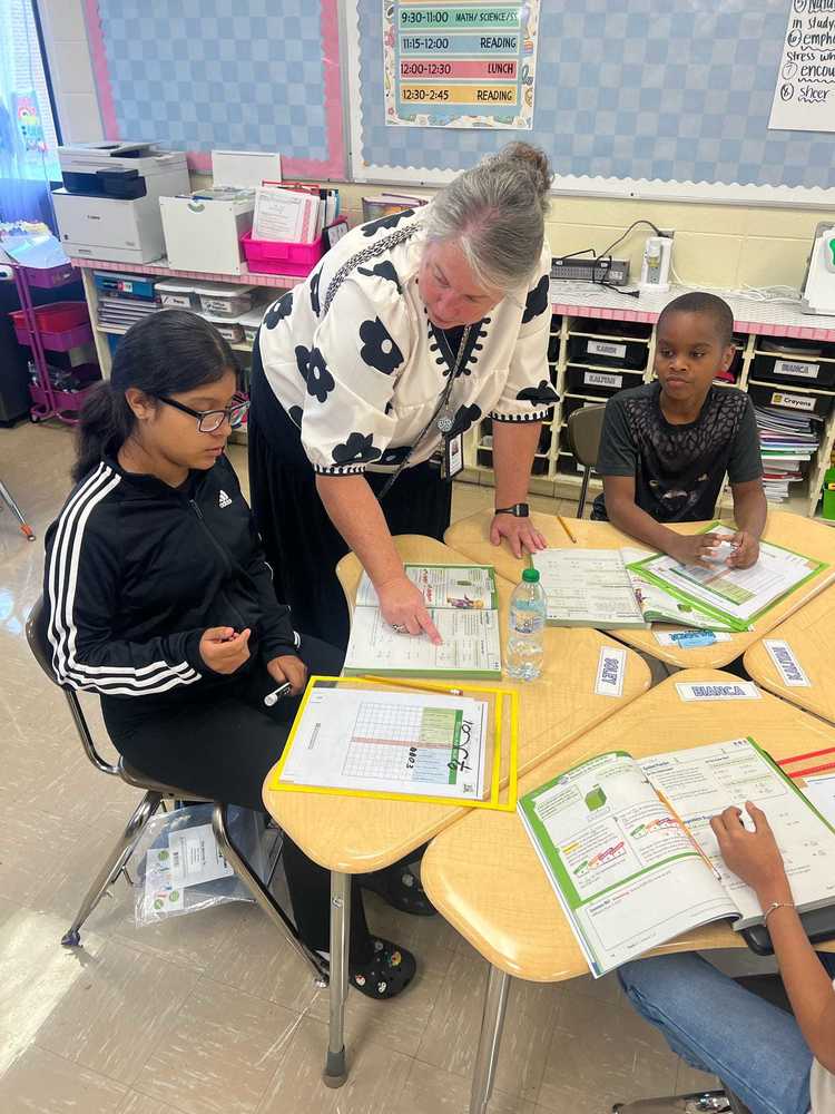 Mrs. West, our math coach, is standing over the table with two students at a desk, pointing to math problems in a notebook and explaining problem-solving techniques. The students are watching attentively and engaging with the lesson.