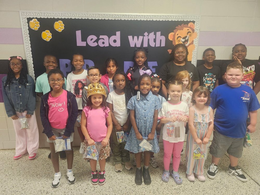 A group of 18 students, ranging from Kindergarten to 5th grade are posed in front of a bulletin board that says "Lead with PRIDE"