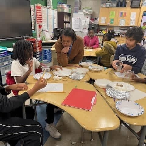 Students pictured at the table with Mrs. Steele -gate teacher while students have a plate and cup with water inside