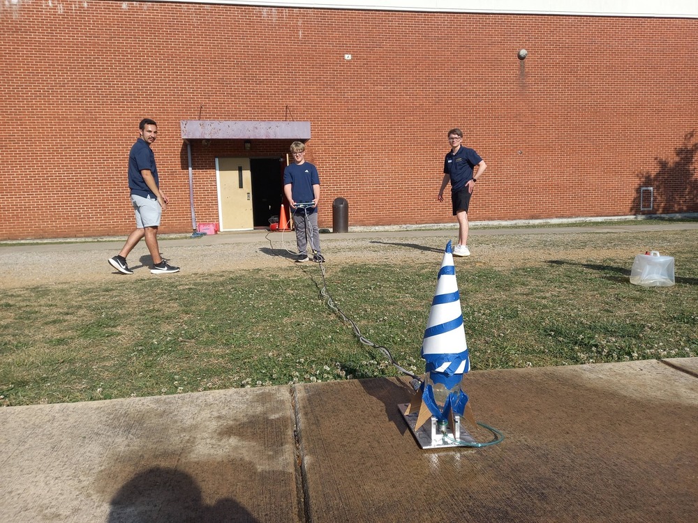 A rocket is in the front and a student is behind it ready to launch it. Two UAH college students are off to the side watching.