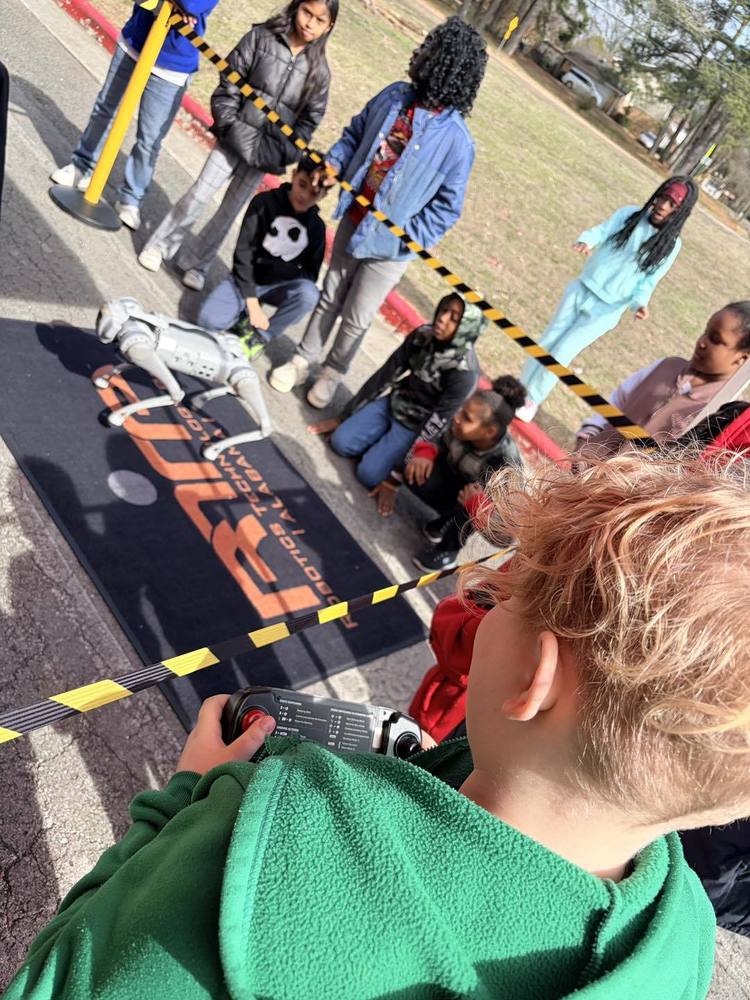 A group of students are standing around outside a mat with a robotic dog. 