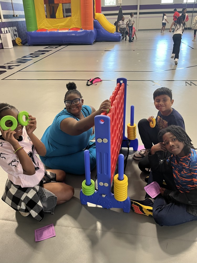 4 students are playing a giant Connect 4 game in the center of the gymnasium. There is a yellow bounce house in the background with kids running around.