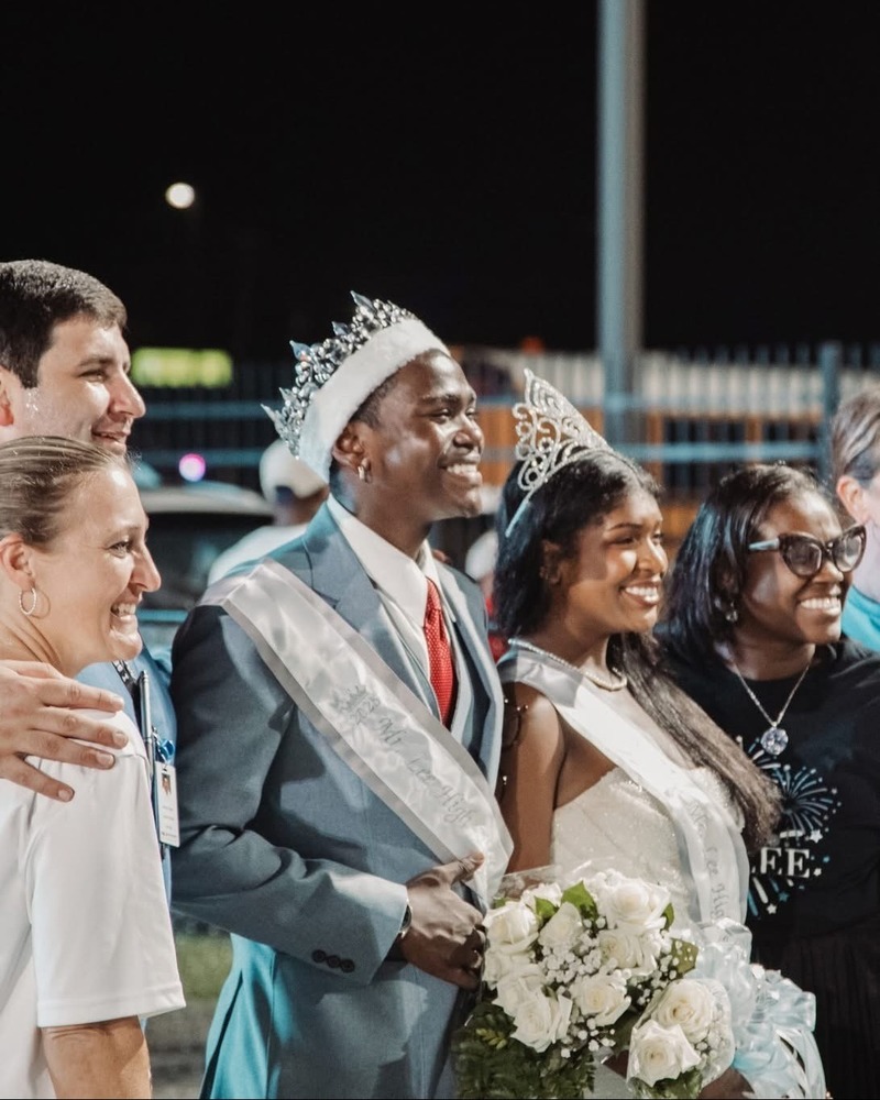 Lee High School Homecoming King Shawn Costley and Homecoming Queen Linda Mathis stand together wearing crowns and sashes, smiling and holding a bouquet of white roses. They are joined by Assistant Principal Megan Carroll Chulan, Assistant Principal Patrick Holder, and Principal Kitrise Torain during the 2025 Homecoming celebration.