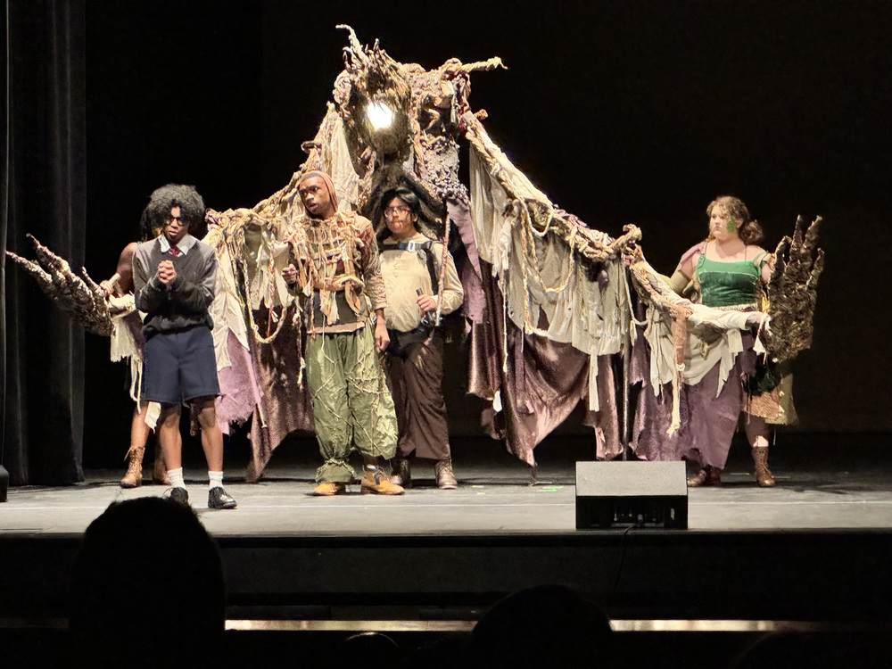 Lee High Theatre students perform a scene from A Monster Calls. Christopher Hart, portraying Conor, stands at the front of the stage beside Christian Bailey as the Monster. Behind them, ensemble members operate a large, tree-like puppet made of twisted branches and draped fabric, creating the illusion of a towering creature.