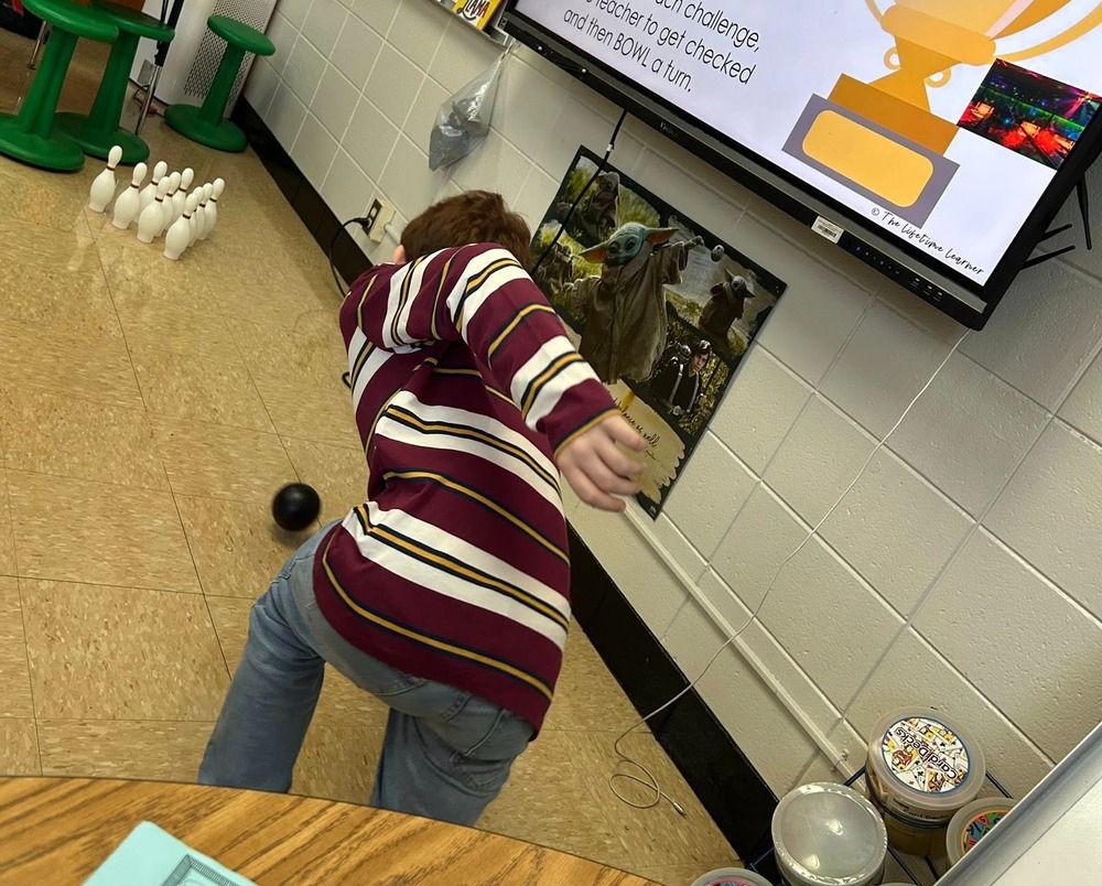 A boy in a red and white striped shirt is in a classroom, rolling a bowling ball towards a set of bowling pins.