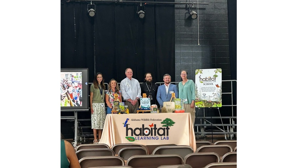 Mrs. Herndon our principal, Mrs. James centeral office representative, three People from Wild Life Federation ,Mr. Avant on a stage in GSES lunchroom with two poster on either side one with Habitate and the other a colorful picture, and a habitat banner on the table Infront of them