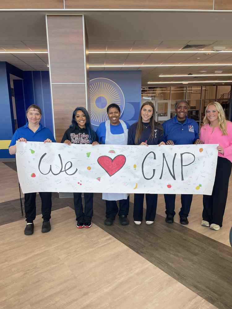 Teaching and Training students pose with child nutrition program workers holding a sign that says We "heart" CNP. The sign has multiple signatures and messages of thanks, as well as, pictures of random fruit.