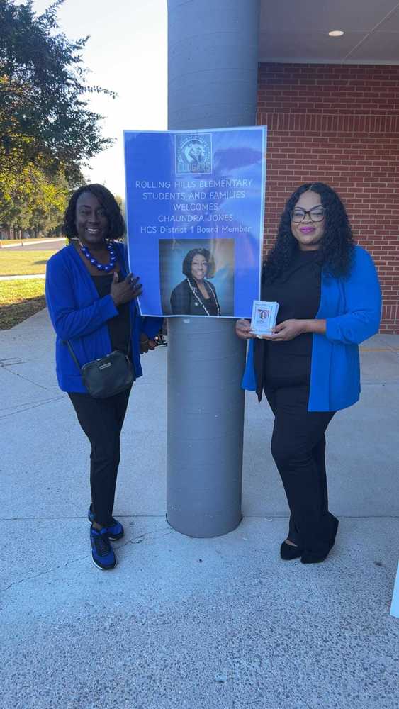 Principal Agun and HCS District 1 Board Member Chaundra Jonesare standing outside near a pillar holding a poster that reads, “Rolling Hills Elementary Students and Families Welcomes Chaundra Jones, HCS District 1 Board Member.” Both women are dressed in black and blue, smiling for the photo.