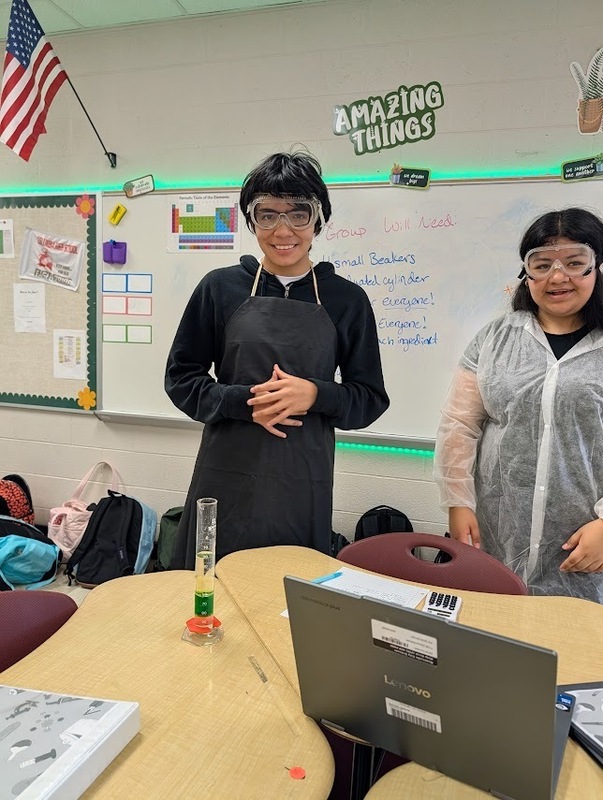 2 students in science class standing in front of desk one wearing a black apron and the other  wearing a white lab jacket both with goggles on