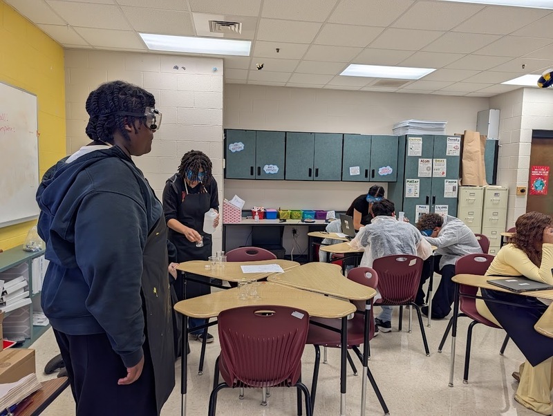  Students in science class standing and sitting around tables wearing jackets, safety glasses using tools collaborate on an assignment or experiment