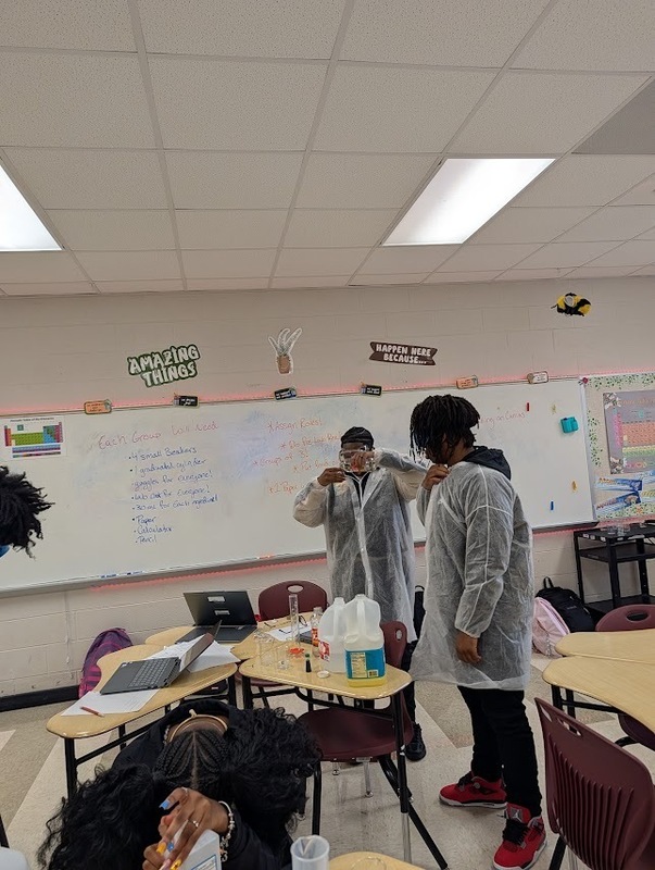 2 students standing in science class wearing white lab jackets and goggles standing in front of white board and desks