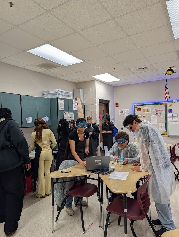  Students in science class standing and sitting around tables wearing jackets, safety glasses using tools collaborate on an assignment or experiment