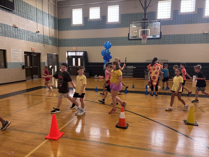 15 4th grade students at the end of the running in the gym doing their cool down lap with blue balloons in the background