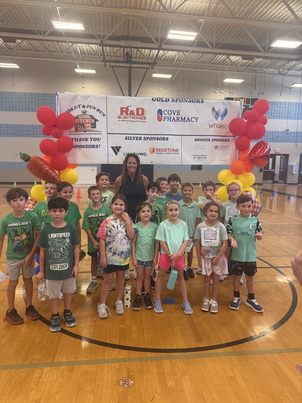 Miss Yarbrough's second grade picture in front of the sponsor banner with yellow and red balloons on each side in the gym celebrating their run during boosterthon