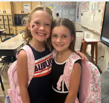 Two girls in Auburn cheering outfit both with same color backpacks one has an A U on her check for college day for college and career week. They are in a classroom at school.