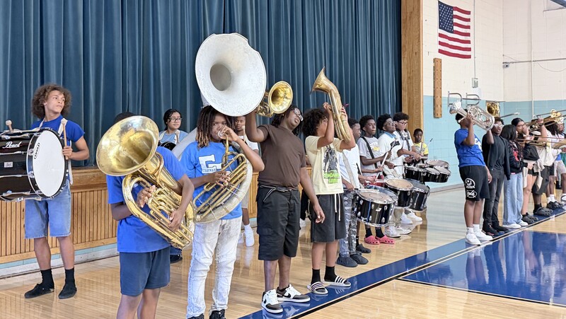 Members of the school band lined up on the basketball court playing brass instruments, including sousaphone and tubas, and percussion drums in front of blue stage curtains.
