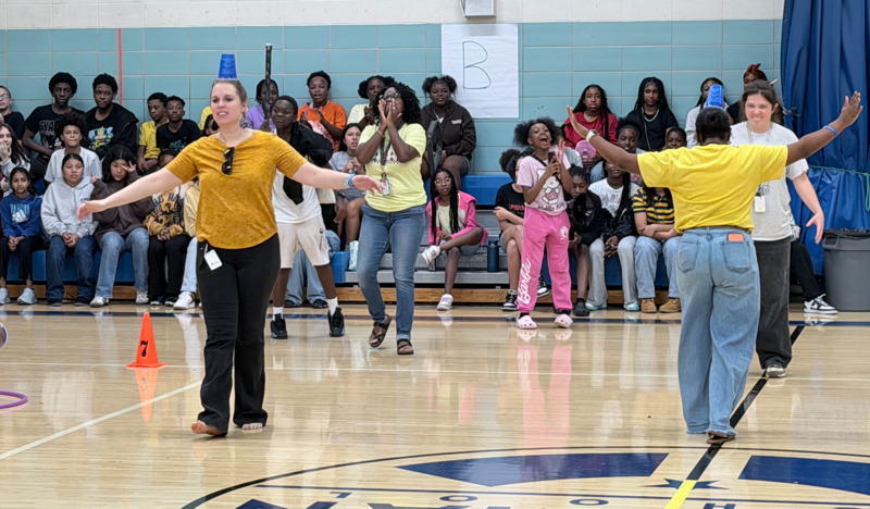 Staff and students compete in a balance game on the basketball court with plastic cups on their heads as spectators in the bleachers cheer.