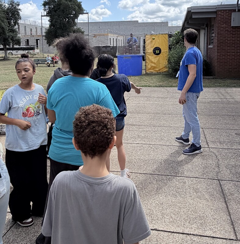 A line of students waiting on a paved walkway outside to throw a ball at a yellow target trigger on a blue dunk tank holding Mr. Thornton.
