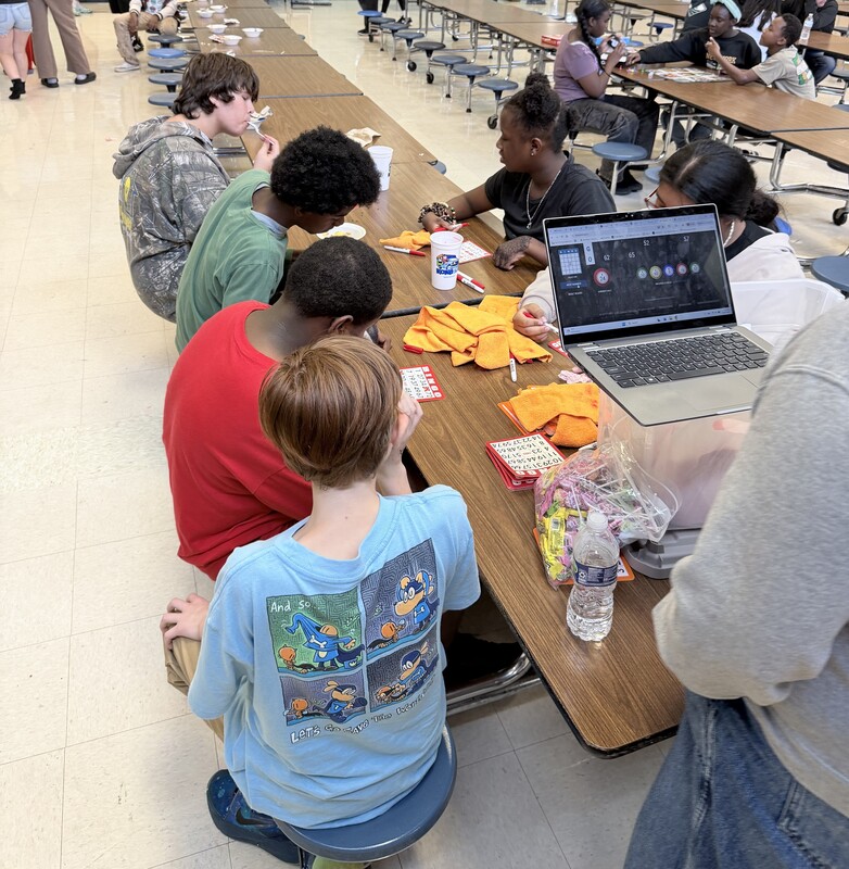 Students sit along long cafeteria tables playing Bingo using red game cards with a laptop displaying game numbers nearby.