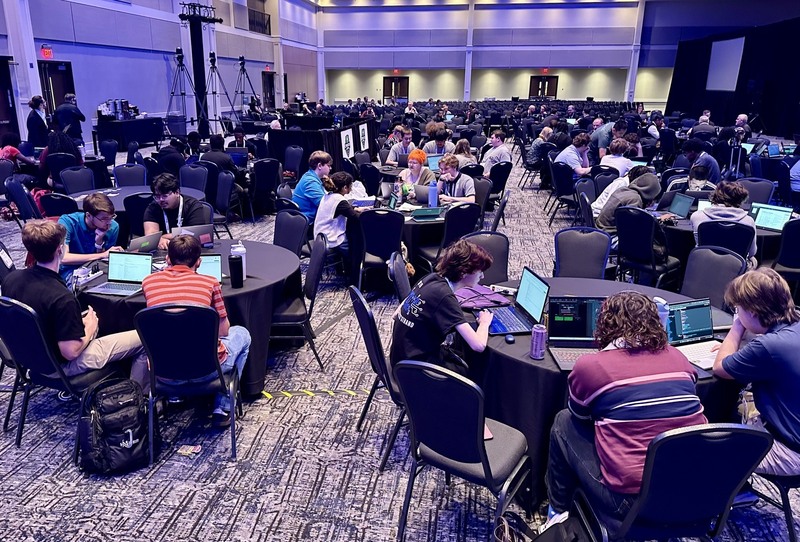 Students sit in groups at the Cyber Summit while working on their laptops.