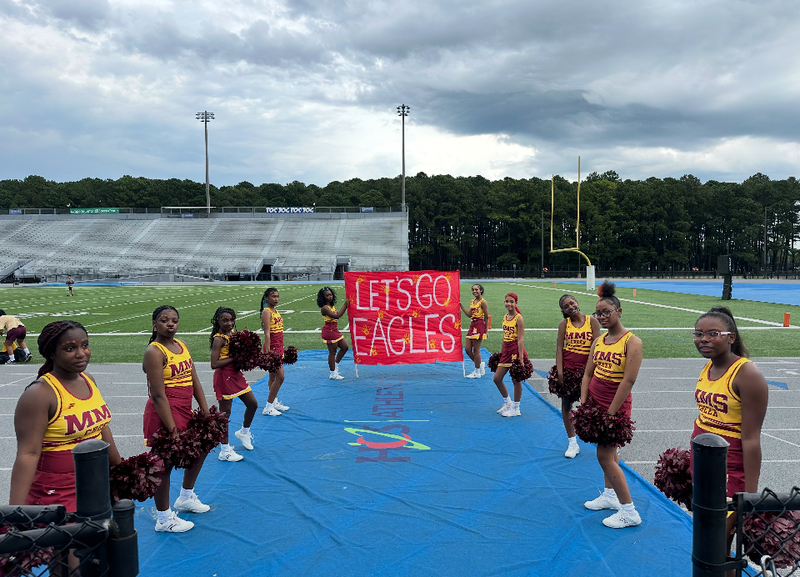 Cheerleaders in crimson and gold uniforms flank a red sign reading "Let's Go Eagles!"