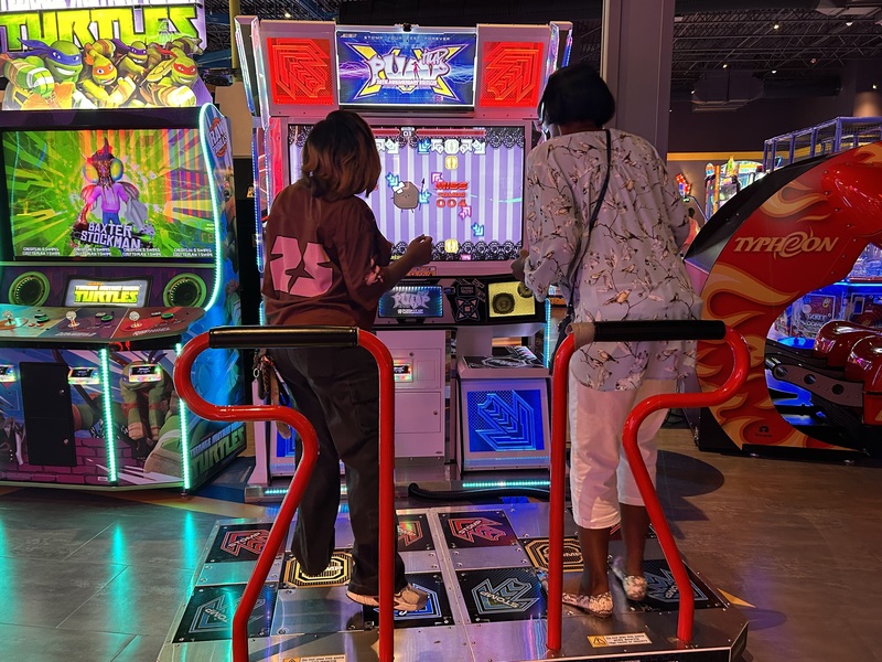 Ms. Gray and Ms. Morrow are actively engaged in a dancing competition video game at the Main Event.