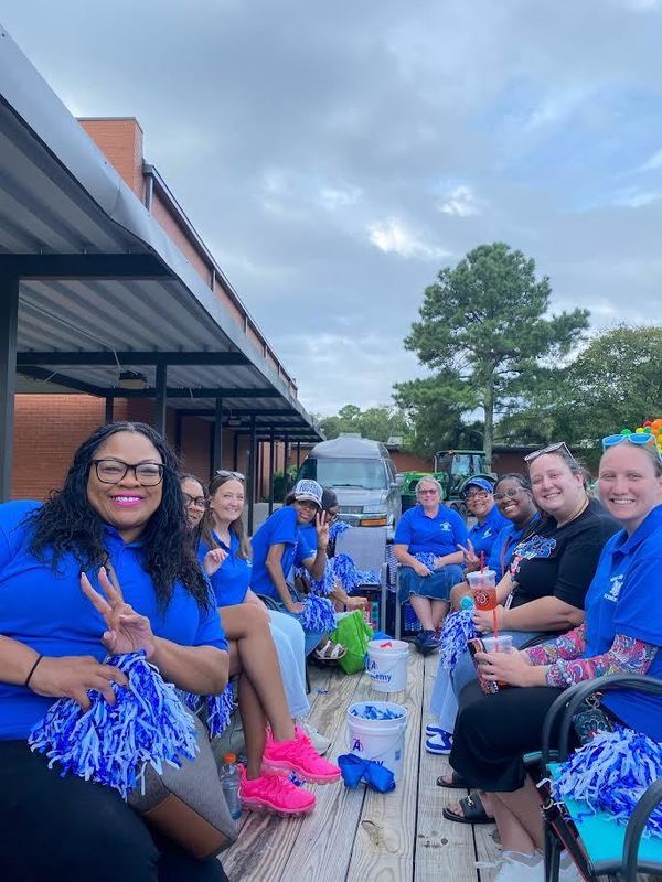 Principal Agun and RHES staff members sit together on a parade float, smiling and holding blue pom-poms. Principal Agun is in the front left making a peace sign, and the group is dressed in matching blue shirts, showing school spirit.